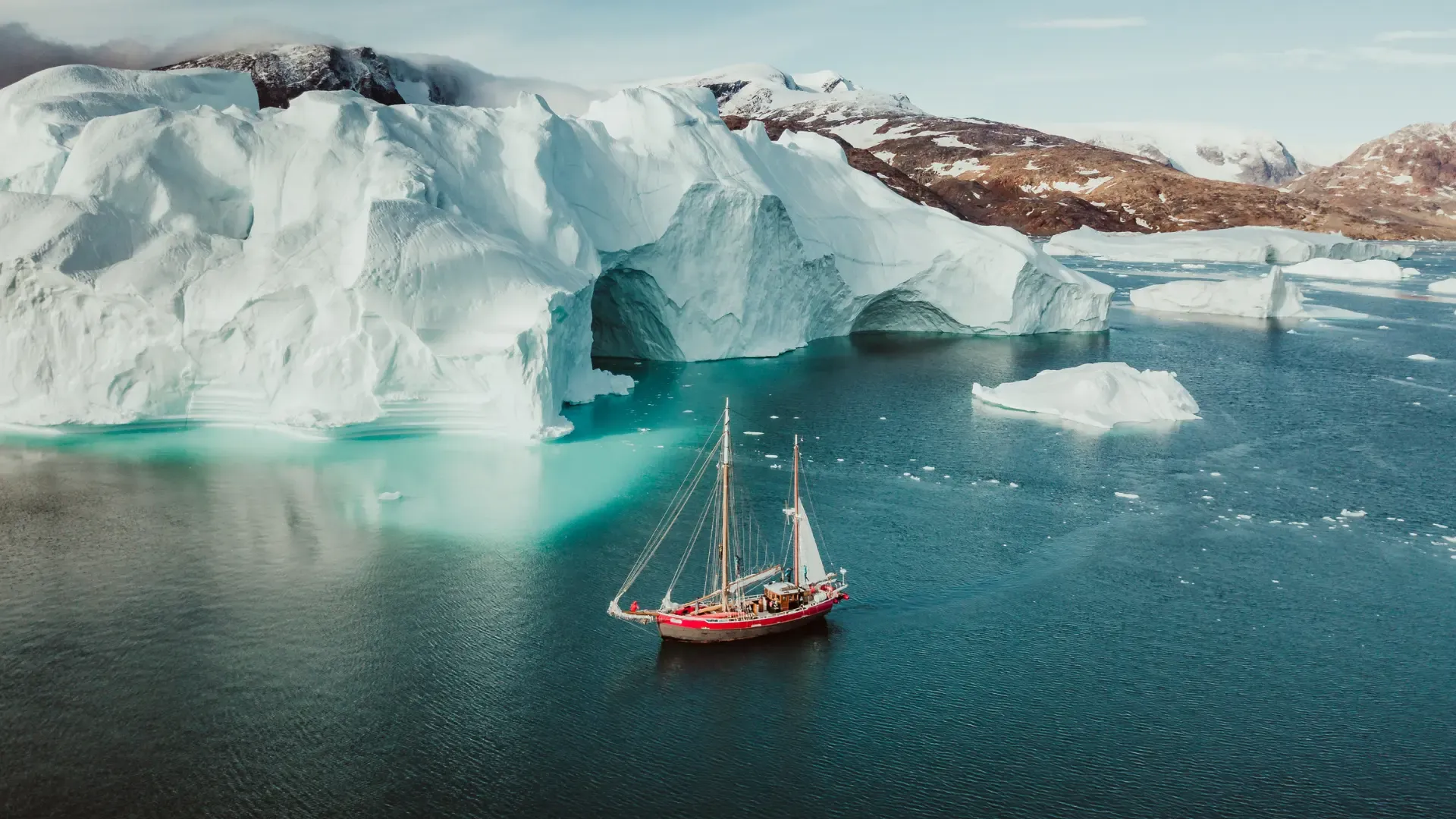 A red sailing ship beneath a towering arched iceberg in turquoise Arctic water
